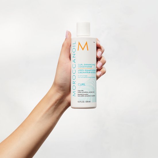 Een hand houdt een witte fles Moroccanoil Curl Enhancing Conditioner (250 ml) vast. Fles met oranje M logo.