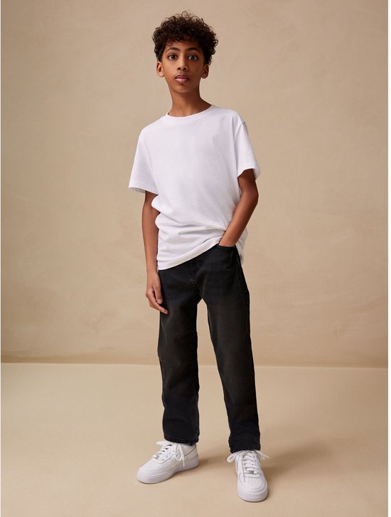 Jongen met krullend haar, witte T-shirt, zwarte spijkerbroek en witte sneakers, staand.