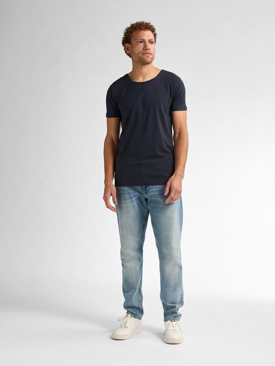 Man met krullend rood haar, sproeten, donkerblauw t-shirt en lichte spijkerbroek.