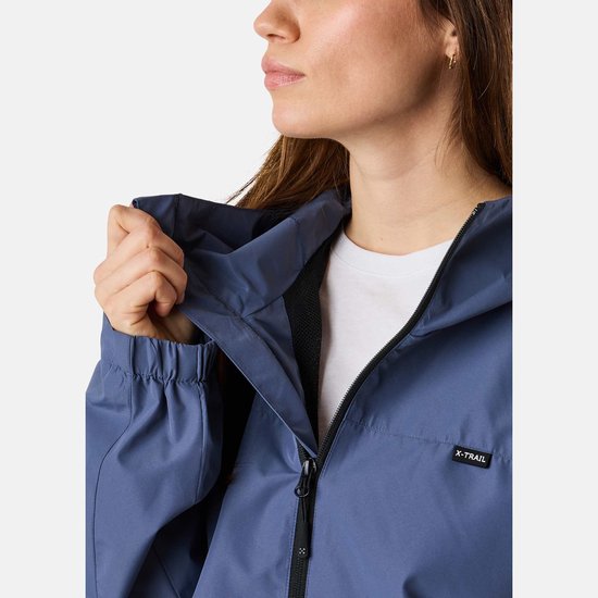Close-up van een vrouw die het opvouwbare deel van de capuchon van een blauw X-Trail jack vastpakt. Zwart X-TRAIL logo op de borst.