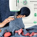 Handen van een persoon die naait aan een blauwe stof op een witte Rynix naaimachine.