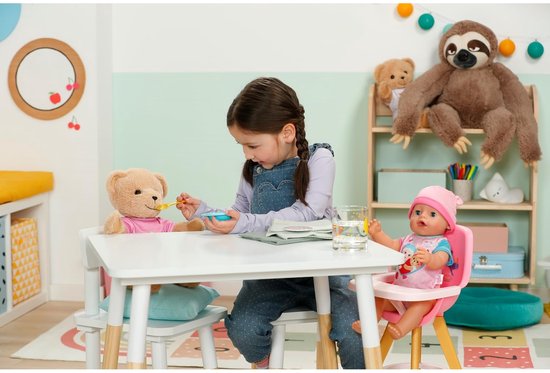 Meisje voert een teddybeer en een baby in een kinderstoel aan een tafel.