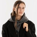 Een vrouw met lang bruin haar, die een donkere jas en een grijze gebreide colsjaal met capuchon draagt.