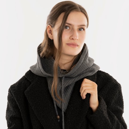 Een vrouw met lang bruin haar, die een donkere jas en een grijze gebreide colsjaal met capuchon draagt.