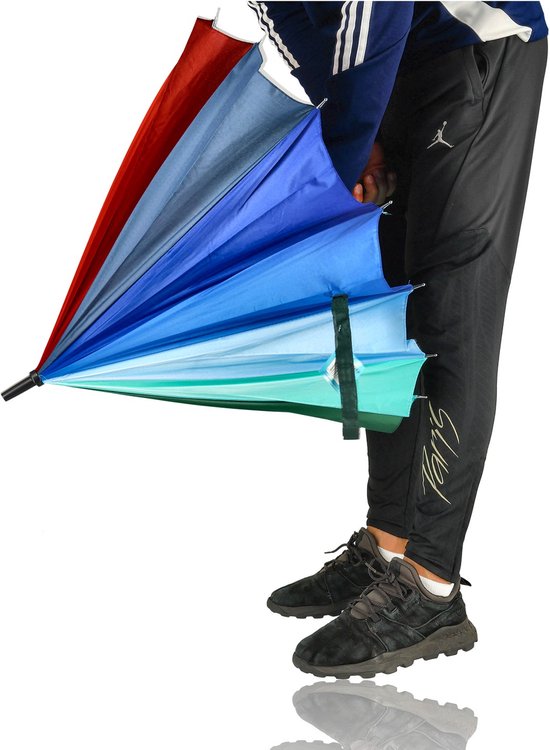 Een persoon met sportbroek kijkt naar een gedeeltelijk gesloten regenboogparaplu met veelkleurige vlakken in blauw, groen en rood.