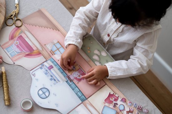 Meisje plakt stickers op een kartonnen speelset die een poppenhuis voorstelt.