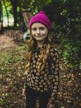 Meisje met lange rode krullen en sproeten, draagt een zwart shirt met bloemenprint en een roze muts.