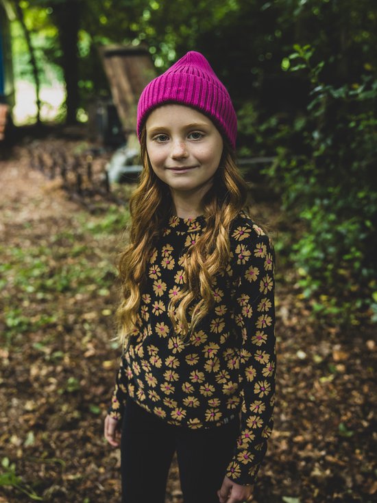 Meisje met lange rode krullen en sproeten, draagt een zwart shirt met bloemenprint en een roze muts.