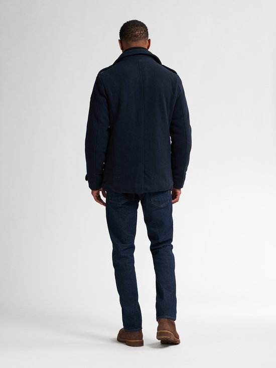 Man staand op rug gezien, met donkerblauwe peacoat, jeans en bruine schoenen.