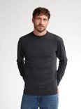 Man staat frontaal in een donkergrijze gebreide trui en blauwe jeans.