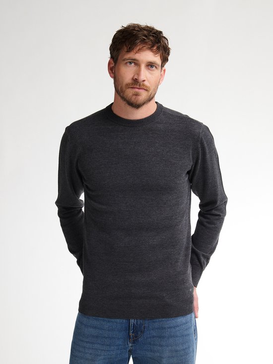 Man staat frontaal in een donkergrijze gebreide trui en blauwe jeans.