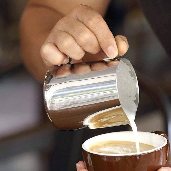 Hand schenkt melk uit een klein roestvrijstalen kannetje in een bruine mok met latte art.