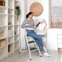 Een vrouw met een gestreepte trui en spijkerbroek zit op een witte opvouwbare trap en leest een boek.