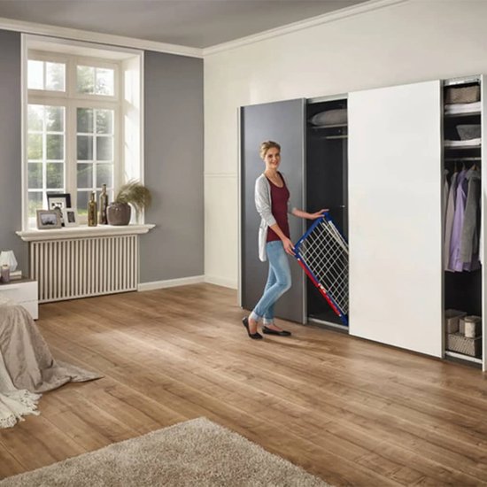 Vrouw met grijze vest en spijkerbroek houdt een opgevouwen wit droogrek vast bij een open kastenwand.
