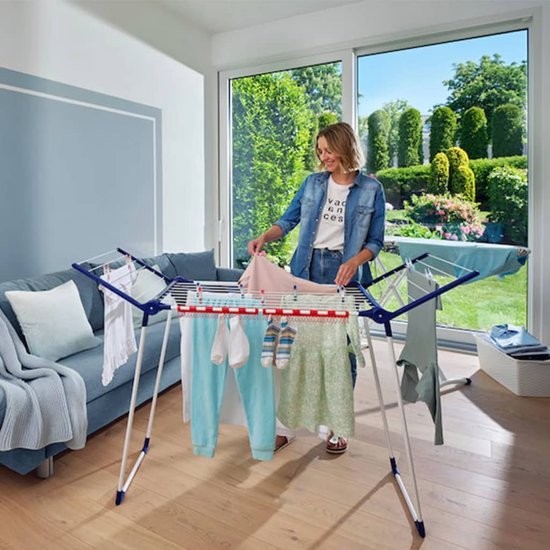 Vrouw in spijkerbroek en spijkerjack sorteert kleding op een wit droogrek in een modern ingerichte woonkamer.