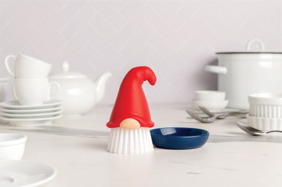 Een afwasborstel in gnome-vorm met rode puntmuts en beige neus, witte borstelharen, in een donkerblauwe houder, naast een omgekeerde blauwe schaal.