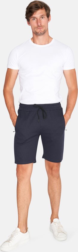 Man staand met witte t-shirt en donkerblauwe korte broek, met trekkoord en ritssluitingen aan de zijkant.