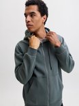 Model trekt aan de kraag van een donkergroene hoodie met ritssluiting.