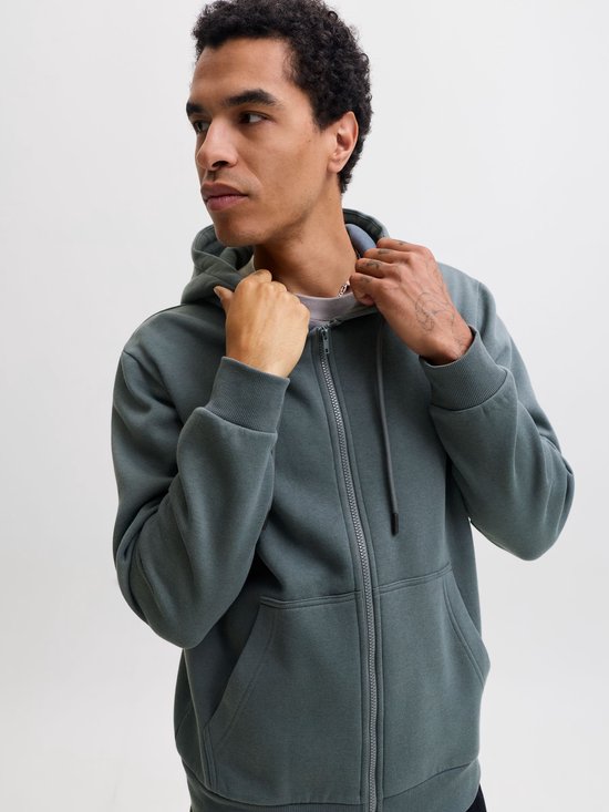 Model trekt aan de kraag van een donkergroene hoodie met ritssluiting.