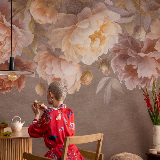 Behang met een bruine achtergrond en grote, zachte bloemen in pastelkleuren. Een vrouw in een rode kimono zit aan tafel met een kopje.