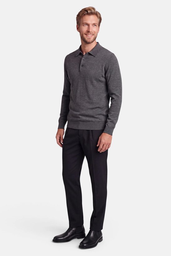 Lachende blonde man in een donkergrijze polotrui, donkere pantalon en zwarte schoenen.