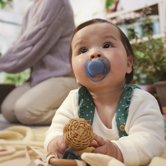 Baby met blauwe fopspeen in de mond, blauwe rand rond het fopspeen, liggend op een geel kleed.