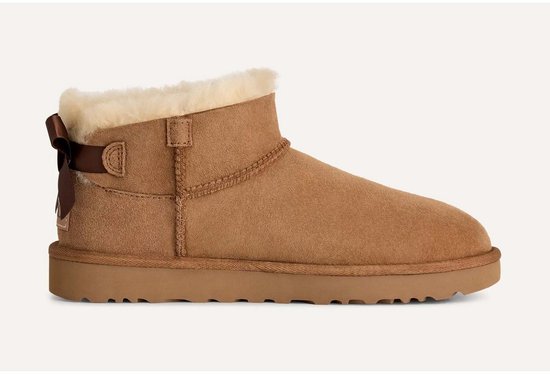 Zijaanzicht van een cognackleurige suède UGG Ultra Mini laars met crèmekleurige voering en strik aan de achterkant.