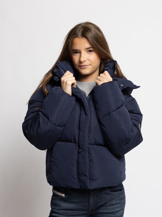 Meisje met lang bruin haar in een donkerblauwe pufferjas die de kraag vasthoudt, met een grijze top eronder.