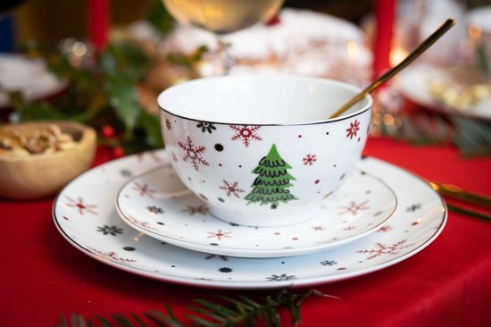 Stapel van een wit porseleinen bord en kom met kerstboom- en sneeuwvlokprint op een rode kersttafel.
