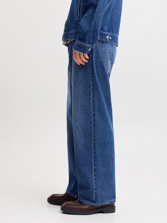 Man met korte blonde haren, donkerrode pet, jeansjack en wijde jeans, staand opzij.