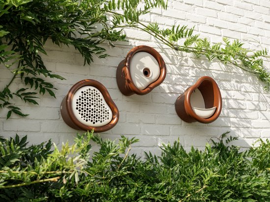 Drie bruine en crème vogelhuisjes op een witte bakstenen muur. Van links naar rechts: één met rooster, één met ronde opening, één met schaaltje. Groene planten aan de zijkanten.