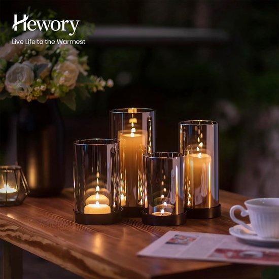 Vier glazen windlichten met brandende kaarsen op een houten tafel naast een vaas met bloemen.