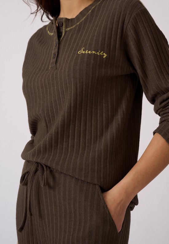 Model in bruine pyjamaset van ribtricot. De tekst "Serenity" staat geborduurd op de borst. De pyjamabroek heeft een strikceintuur.