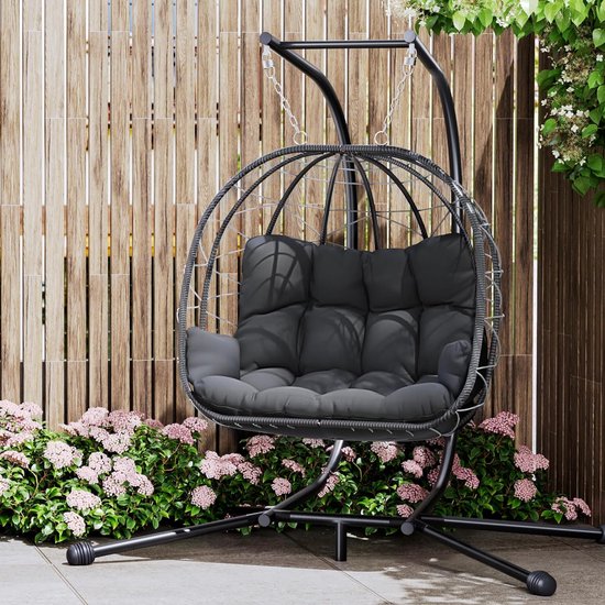 Grijze hangstoel in rotanlook met dik, grijs kussen, staande op een zwart metalen onderstel, met houten schutting op achtergrond.