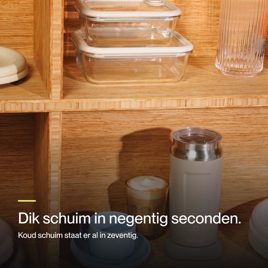 Beige melkopschuimer met zilveren deksel en knop, naast twee glazen met koffie en melkschuim.