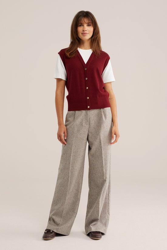 Model in een gebreide bordeauxrode vest met knopen over een wit t-shirt, met een gemêleerde grijze wijde broek.