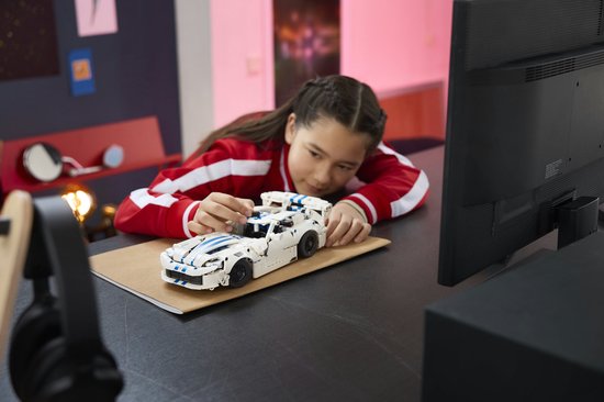 Een meisje met een rode jas legt een witte LEGO Technic Dodge Viper GTS-R sportauto met blauwe strepen op een bureau.
