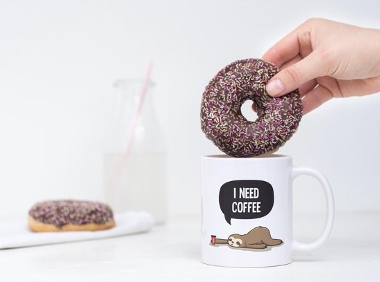 Tasse blanche avec texte 'I NEED COFFEE' et paresseux, tandis qu'un beignet est placé dans la tasse.