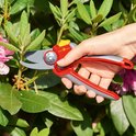Grijze en rode snoeischaar met softinleg, in een hand gehouden bij roze bloesems en groene bladeren.