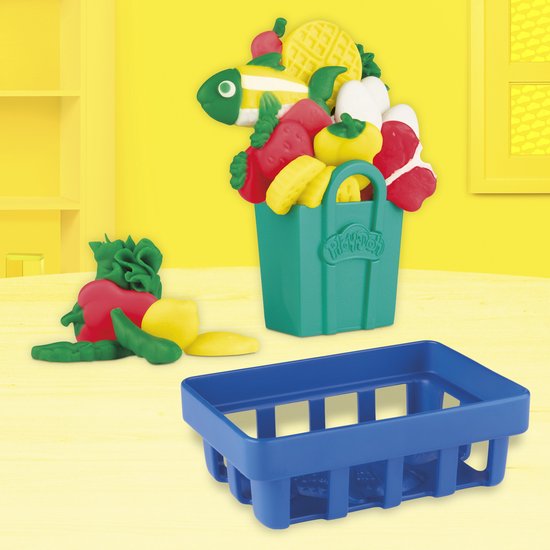 Een groene mand vol speelgoedfruit en -groenten, met een blauw winkelmandje en extra groenten op een gele tafel.