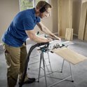 Man met blauwe polo en beige werkbroek zaagt een houten plank met een cirkelzaag op een geleiderail.