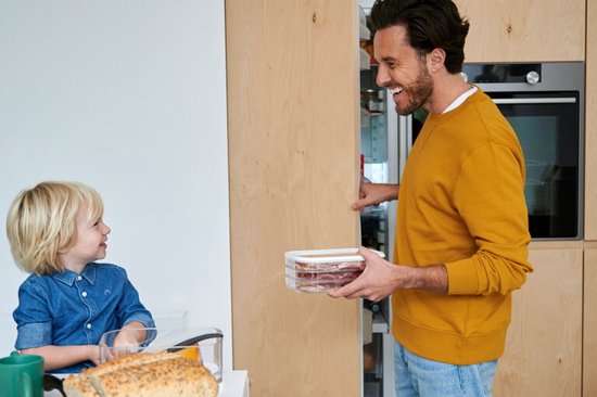 Een man opent een koelkast en houdt een Mepal bewaardoos met vleeswaren vast, kijkend naar een jong kind.