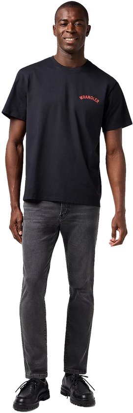 Man draagt een zwart t-shirt met rode Wrangler opdruk en grijze jeans. Zwarte schoenen.