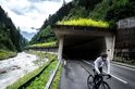 Man op racefiets onder een betonnen tunnel met groen dak, rijdt langs een rivier in een witte fietsregenjas.