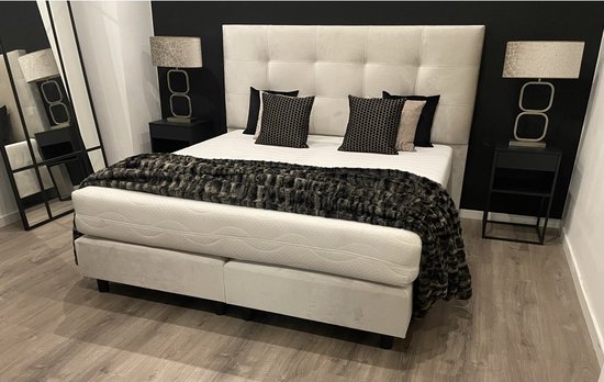 Moderne slaapkamer met beige boxspring, vier kussens, sprei, twee nachtkastjes met lampen en een grote spiegel.
