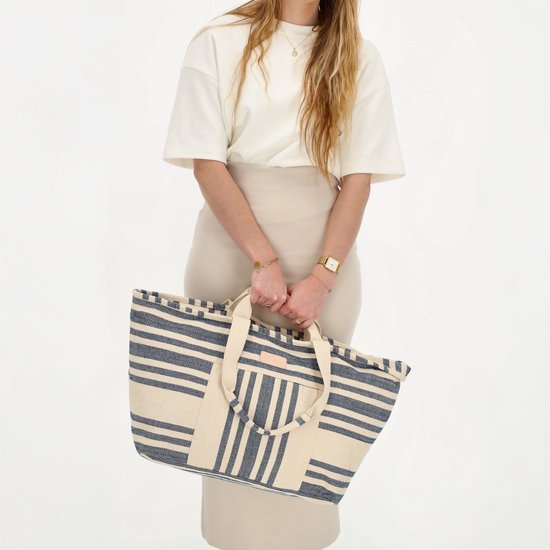 Vrouw met lange blonde haren in beige kleding, draagt een grote gestreepte strandtas met beige hengsels.