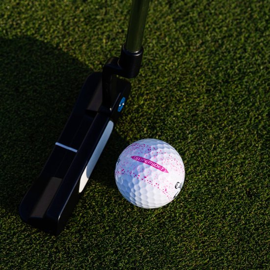 Callaway golfbal met roze spetters en Callaway logo naast een zwarte golfputter op een groen grasveld.
