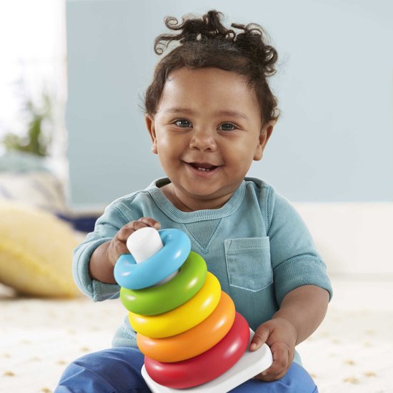 Een baby met krullend haar lacht en houdt een Fisher-Price stapelpiramide vast met vijf gekleurde ringen. De bovenste ring is lichtblauw.