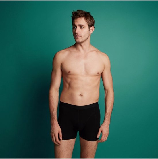 Man in zwarte boxesshorts, zittend voor een groene achtergrond.