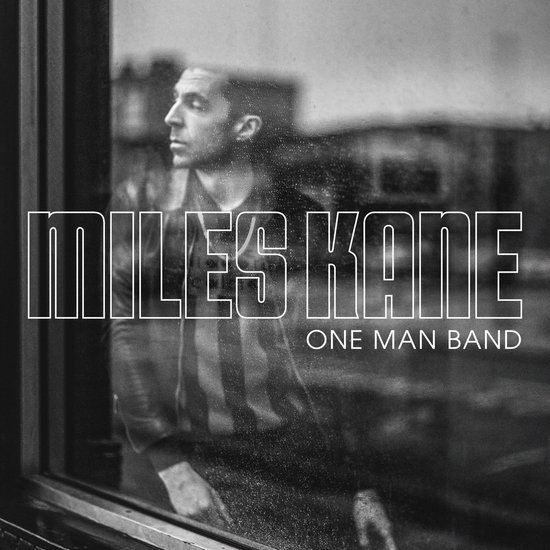 Miles Kane: One Man Band. Een zwart-wit afbeelding van Miles Kane die door een raam kijkt, gekleed in een jas.